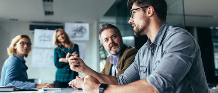 Vier Personen in einem modernen Besprechungsraum diskutieren konzentriert. Ein Mann im Vordergrund erklärt etwas mit einem Stift in der Hand, an der Wand hängen technische Zeichnungen.
