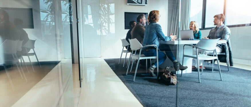 Fünf Personen sitzen in einem modernen Konferenzraum an einem Glastisch und führen eine Besprechung. Laptops und Notizen liegen auf dem Tisch, Sonnenlicht fällt durch große Fenster.