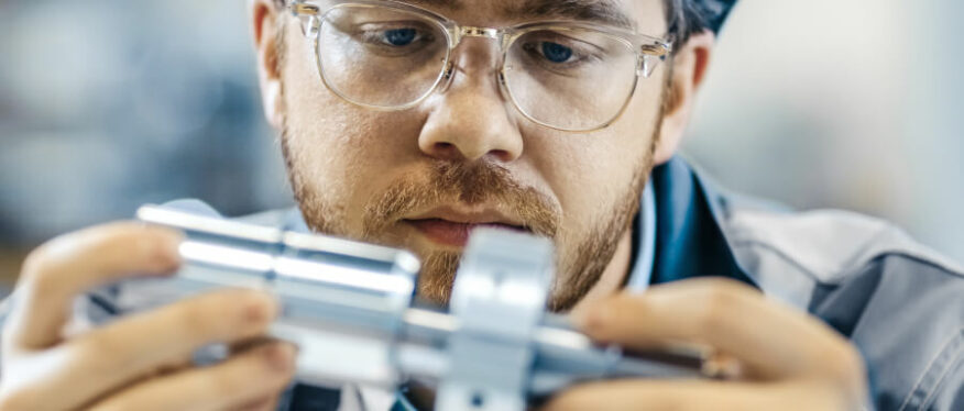 Mitarbeiter mit Schutzbrille und Arbeitskleidung inspiziert konzentriert ein präzise gefertigtes Metallbauteil in der Hand.