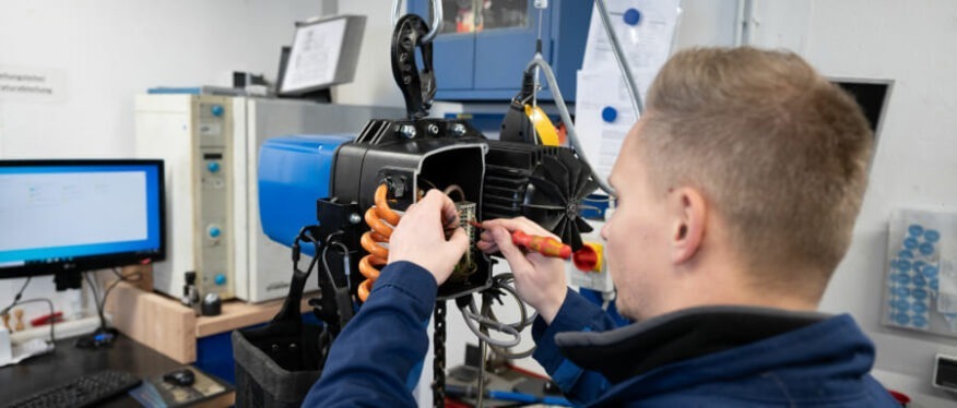 Servicetechniker führt Wartungsarbeiten an einem geöffneten Elektrokettenzug mit blauem Gehäuse in einer Werkstatt durch. Im Hintergrund sind Prüfgeräte und ein PC sichtbar.